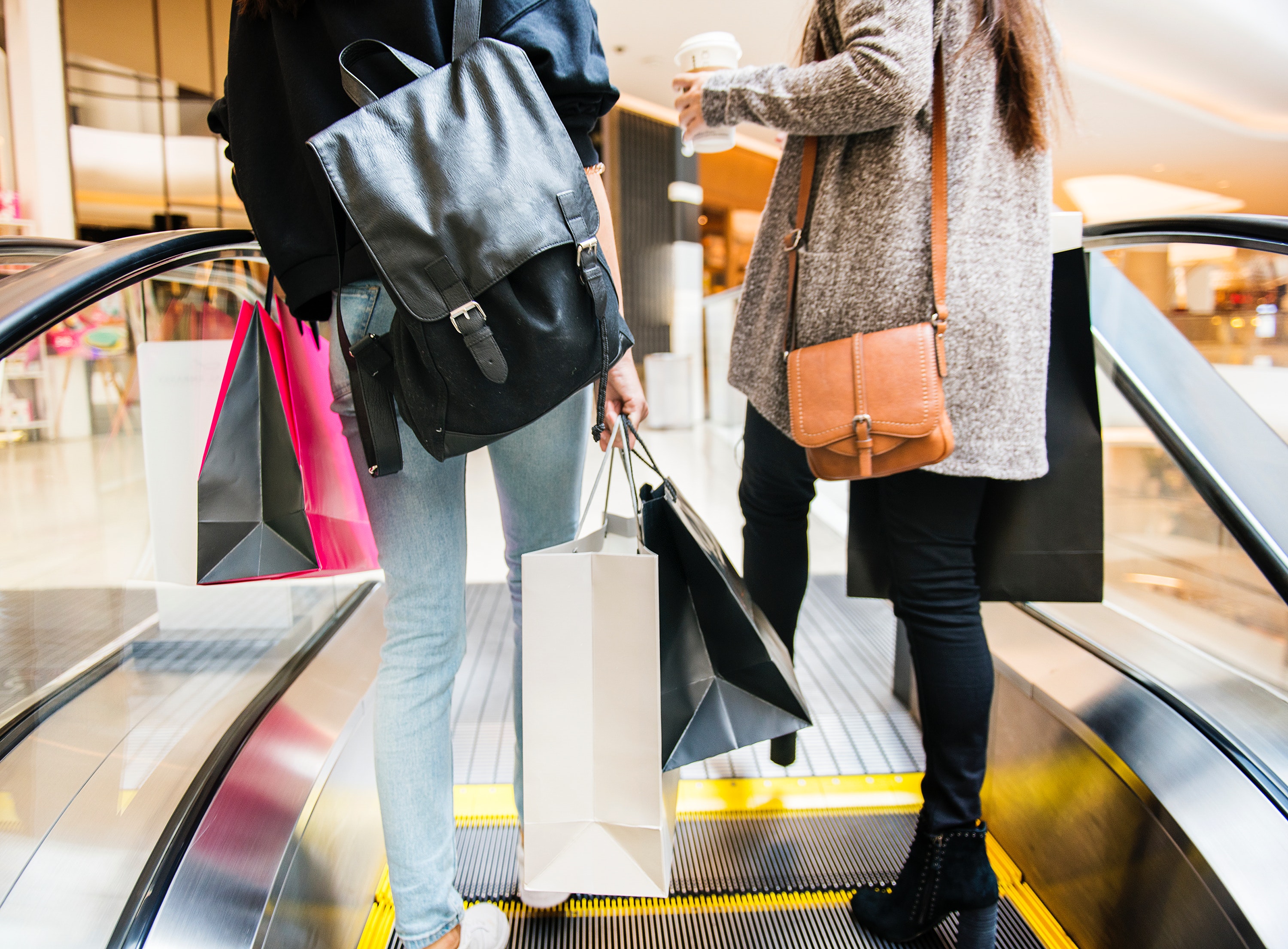 Two women holding shopping bags while riding an escalator.
