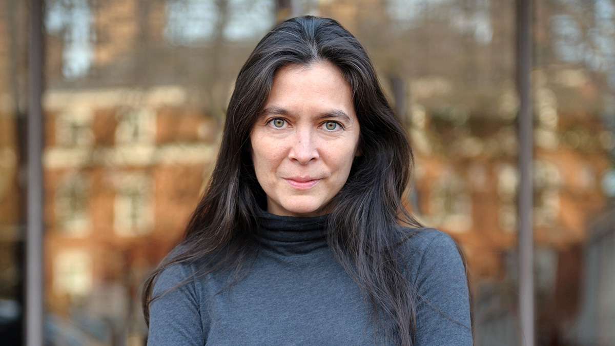 Diane Paulus wearing a gray turtleneck in front of a reflective surface in a metropolitan area.